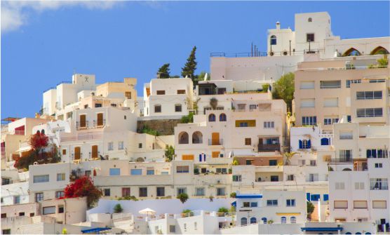 Vista de edificios y casas blancas con muchas ventanas acristaladas y con persianas azul marino, amarillo y tonos naranjas. Todas ellas tienen terrazas en sus azoteas. Entre ellas se ven árboles, plantas y enredaderas con flores de color fucsia. Los edificios están sobre una colina. La imagen ha sido tomada desde abajo en un día soleado, resaltando el cielo azul celeste con una nube blanca.