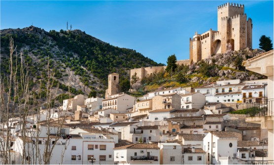 Imagen de uno de los pueblos blancos de Almería, Velez Blanco, con un imponte castillo en lo alto de una colina y a modo de cascada las casas blancas. Al fondo podemos ver otra colina rocas y arbusto.
            
