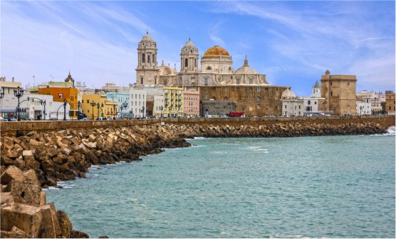 Vista panorámica de la catedral de Cádiz que se eleva de forma monumental a orillas del mismo golfo de Cádiz. Bordeando la costa hay un enorme dique formado por grandes bloques de piedra y sobre este una calle con vehículos transitando, semáforos y farolas de hierro forjado. Junto a ella se encuentra la catedral y viviendas de diferentes colores.