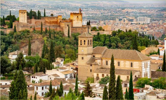 Vista panorámica de la ciudad de Granada desde lo alto de una colina desde donde se divisa la Alhambra rodeada por una arboleda. A sus pies se encuentra gran parte de la ciudad y los emblemáticos barrios de Albaicín y Sacromonte. 