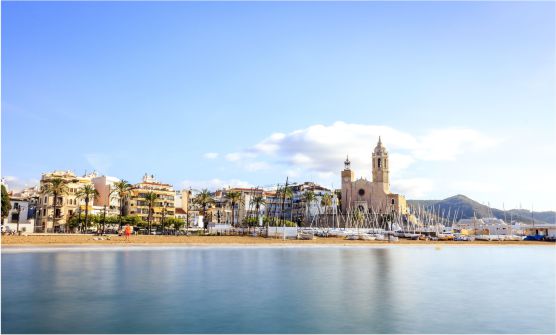 Vista panorámica de la localidad de Sitges tomada desde el mar, desde donde se contempla la playa en una mañana con mucha calma, pequeñas embarcaciones de recreo sobre la arena, viviendas al fondo y la iglesia de San Bartolomé y Santa Tecla situada sobre una pequeña atalaya frente al mar. Detrás de la iglesia podemos ver algunas montañas bajas. 