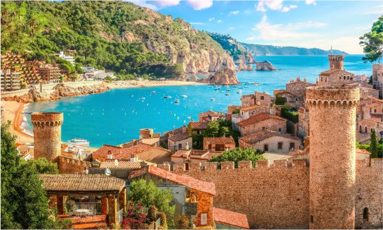 Hermosa imagen del pueblo de Tossa de Mar tomada desde lo alto. Podemos ver el recinto amurallado que protege el centro de la ciudad, así como los tejados y parcialmente las fachadas de las viviendas edificadas sobre una pequeña colina con vistas a la playa y la costa. El mar está salpicado por pequeñas embarcaciones y multitud de boyas, mayoritariamente de color amarillo, delimitan la zona de baño. 