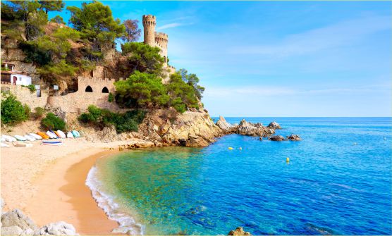 Imagen del Castillo de Lloret de Mar tomada desde la playa. Desde aquí podemos ver pequeñas embarcaciones sobre la arena. Al final de la playa y sobre un montículo al límite de la costa, una escalera sube hacia el Castillo del que podemos ver parte de la fortificación, una torre y una almena. Varios árboles y matas crecen junto al castillo. El mar tiene diferentes tonalidades de azul y turquesa. 