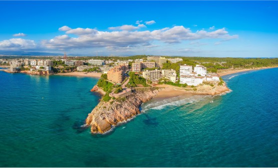 Vista panorámica de la costa de Salou. En la imagen se ven dos playas separadas por un peñón y en ambos lados se pueden ver diferentes edificaciones situadas muy cerca de la costa. El mar tiene diferentes tonalidades de azul que cambia según el reflejo del sol. Detrás de los edificios hay una zona verde salpicada por algunas casas. Algunas nubes blancas y grises decoran el cielo azul. 