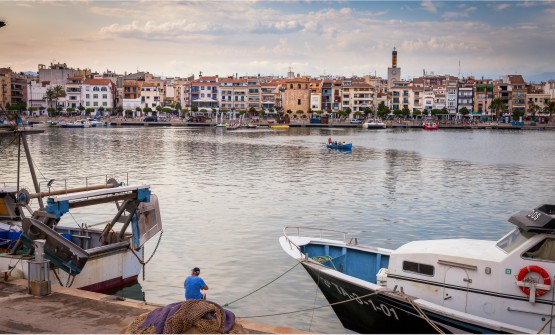 Vista panorámica del puerto de Cambrils. La imagen ha sido tomada desde el puerto pesquero, donde se ve un hombre sentado sobre un bolardo de amarre y junto a una montaña de redes de pesca. En cada lado hay dos barcas grandes de pesca, de una de ellas se ve la proa y de la otra la popa en un mar plateado y en calma. Al fondo de la imagen vemos una serie de edificaciones que bordean el puerto y el campanario de una iglesia que sobresale de entre las viviendas, así como pequeñas barcas amarradas y navegando. 