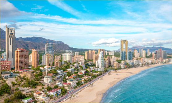 Imagen de la bahía de Benidorm en la que se ven rascacielos de gran altura que despuntan sobre toda la costa. Desde esta panorámica también se puede ver la larga playa de arena blanca bañada por el mar Mediterráneo. 