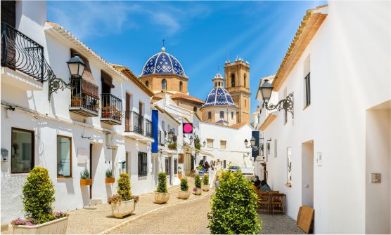 Imagen de una calle peatonal del centro histórico de la ciudad de Altea decorada con jardineras de cemento claro, casas con fachadas de color blanco y balcones y farolas de hierro forjado. Algunos aldeanos se encuentran sentados en las terrazas de la calle. Al fondo de la imagen se puede ver la iglesia de cúpula azul Nuestra Señora de Solace. 