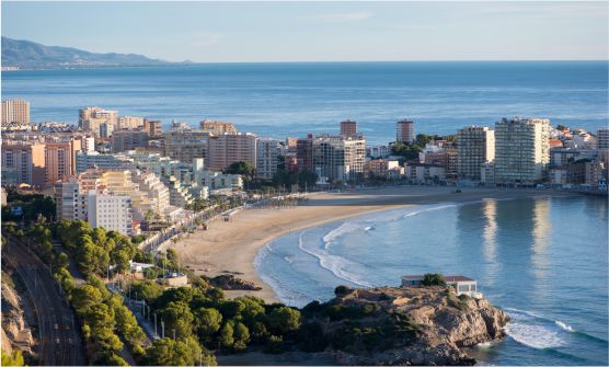 Imagen de la Costa de Oropesa de Mar tomada durante el amanecer, donde se contempla la playa en forma de U, con arena blanca y un mar en calma, sin gente y bordeado por grandes edificios. Al otro lado de la playa, se divisa una gran extensión del mar y la continuación de la costa salpicada por viviendas. 
