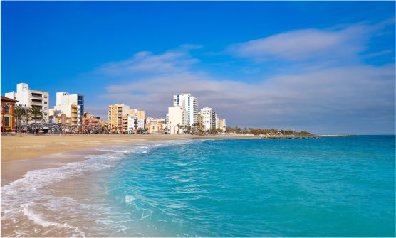 Imagen tomada desde la playa del Forti en Vinaroz en un día claro con algunas nubes al fondo. El mar se agita suavemente en tonos azul turquesa. La espuma de las suaves olas acaricia la arena dorada desde donde se ven edificaciones y casas de media altura. 