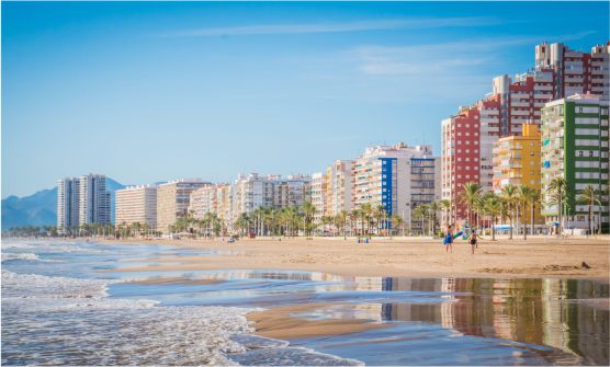 Imagen tomada desde la playa de San Antonio de Cullera desde la que se puede ver como el suave oleaje baña la orilla. Bañistas pasean por la amplia y larga playa desde la que también se ven edifcaciones de gran altura de colores intensos. 
