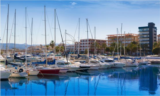 Imagen muy nítida del Puerto de Gandía, con un mar totalmente en calma de color azul eléctrico en el que se reflejan los mástiles de las embarcaciones de recreo amarradas. Al fondo de la imagen se pueden ver unos pocos edificios de diferentes colores. 