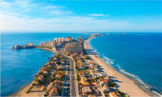 Vista aérea de la Manga del Mar Menor, una larga y estrecha franja de tierra que se prolonga mar adentro. Por el centro de la franja pasa una vía de dos carriles en cada sentido. En ambos laterales de la vía se sitúan multitud de  hoteles y viviendas vacacionales situados hasta el pie de la playa. También se pueden ver varios islotes sin habitar situados a poca distancia de la franja. 