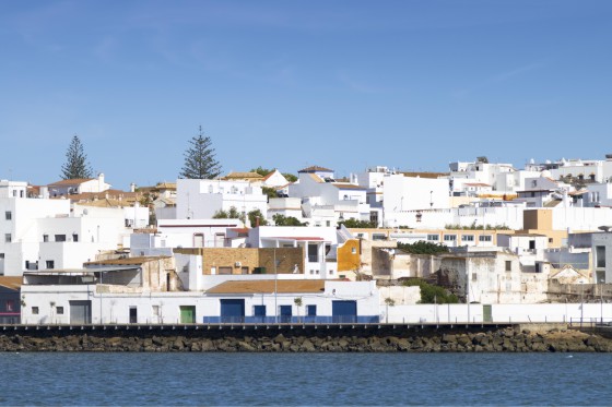 Imagen de la ciudad de Ayamonte tomada desde el río Guadiana, formando una frontera natural entre España y Portugal. Las casas de baja altura están encaladas y tienen tejados planos influenciadas por la arquitectura del norte de África. Frete a ellas está el río y en el fondo de la imagen se ve la copa de dos árboles. 