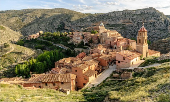Imagen tomada desde lo alto de una colina en la que se puede ver el pintoresco pueblo de Albarracín, con fachadas y tejados en tonos rojizos, destacando los dos campanarios de dos iglesias, uno con tejas en tonos azules y amarillos y el otro con tejas en tonos rojizos y blanco. El pueblo enclavado en un valle con árboles y campos verdes y montañas rocosas. 