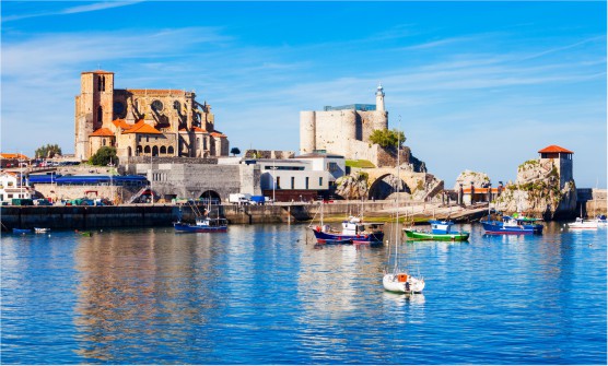 Imagen del puerto de Castro Urdiales en al que se ven algunas barcas pesqueras de color azul y verde intenso, resaltando sobre un mar azul sobre el que se refleja la majestuosa iglesia de Santa María de la Asunción de estilo gótico junto al faro y castillo de Santa Ana. A sus pies un puente medieval testigo de innumerables eventos históricos. 