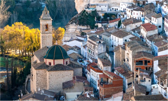 Vista aérea del centro de la localidad de Alcalá del Júcar. Una localidad situada en la ladera de una colina. En el centro se sitúa la iglesia y junto a ella el río rodeado por varios árboles de hojas amarillas. Al fondo están los árboles de las montañas. 