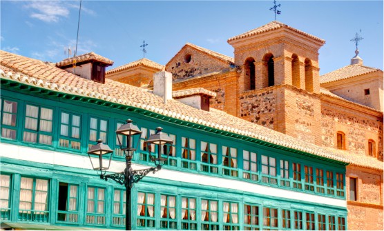 Fachada del Ayuntamiento de Almagro, con dos hileras de ventanas (una inferior y otra superior) con marcos de madera de color verde turquesa. Cada ventana está formada por 6 cristales colocados verticalmente 3 x 3. Algunas de las ventanas tienen las cortinas blancas puestas y otras colocadas para que entre la luz del sol. Al fondo se ve parte de una torre de una iglesia con 2 ventanas verticales en cada cara. 
