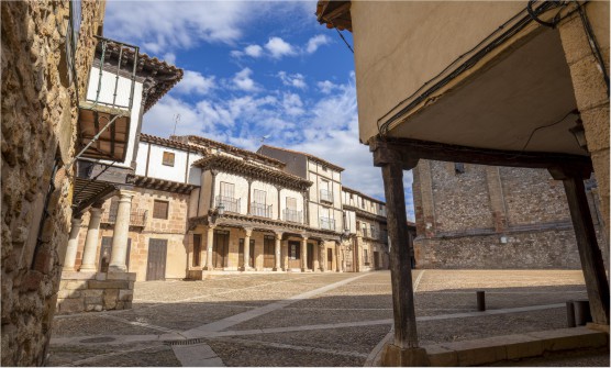 Edificios históricos del casco antiguo de Atienza situados en una plaza empedrada. Algunas de las casas tienen robustas columnas de piedra y vigas de madera que soportan el peso del piso superior. El día es soleado pero cubierto por un manto de nubes. 