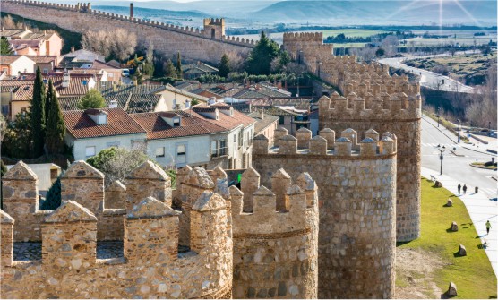 Vista aérea de la ciudad de Ávila en la que se ven parte de las altas murallas que protegen la ciudad. Al fondo de la imagen vemos las montañas con los rayos de sol acariciando las cimas. 