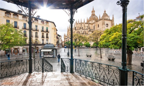 Imagen tomada desde la histórica ciudad de Segovia desde una glorieta con barandillas de hierro forjado situada en una plaza peatonal desde la que se ve la impresionante catedral de Segovia. En la plaza hay personas paseando, un quiosco y un bonito edificio señorial. El suelo está mojado y con pequeños charcos por haber llovido. 