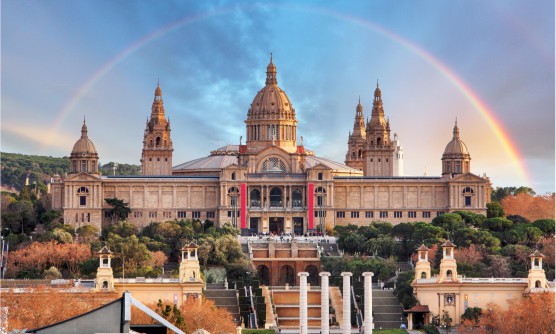 Vista panorámica del Castillo de Montjuïc envuelto por un ligero y casi perfecto arco iris. Un majestuoso y enorme edificio formado por varias torres y una cúpula central. Varias escalinatas dan acceso al edificio rodeado por jardines. 