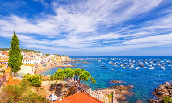 Preciosa imagen panorámica del Calella de Palafrugell. Pintoresca localidad de casas bajas que bordean la playa de la Costa Brava. El mar azul está salpicado por multitud de pequeñas embarcaciones en un día soleado, aunque cubierto por algunas nubes. 