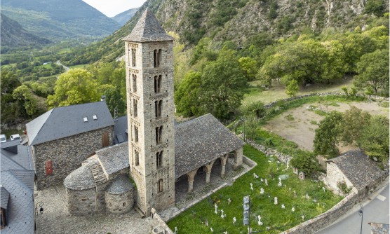 Vista aérea de la Iglesia Románica de Santa Eulalia en la localidad de Erill la Vall, Vall de Boí, con un magnifico campanario de piedra y 2 ventanas verticales en cada una de sus 4 caras y pisos. Los techos de las viviendas que rodean la iglesia son de pizarra y el suelo está empedrado. En uno de los laterales de la iglesia se sitúa un pequeño cementerio del que únicamente se ven las cruces sobre la hierba. 