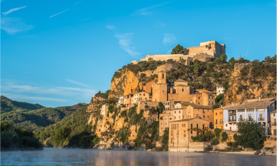 Imagen de la ciudad de Miravet tomada durante el amanecer, una ciudad situada en la misma orilla del río Ebro que crece verticalmente sobre una colina rocosa. Junto a esta colina se extienden una pequeña cordillera tupida de árboles. 