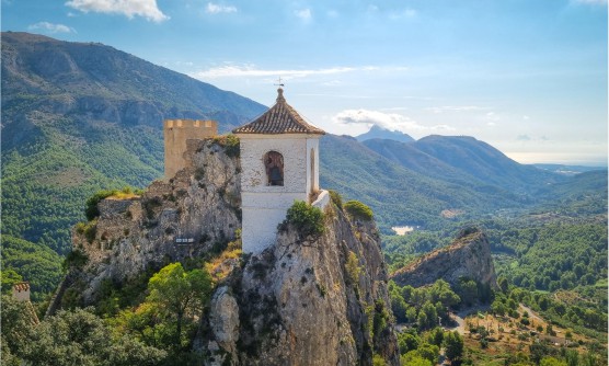 Imagen del Castillo de Guadalest en las tierras altas. En la imagen se ve un campanario que se alza sobre una cumbre montañosa con un enorme precipicio. Detrás del campanario una torre fortificada.  