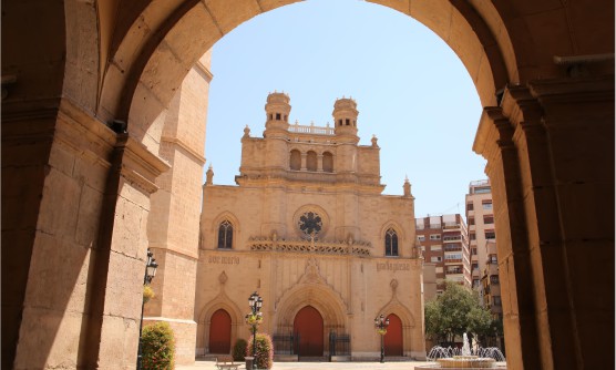 Imagen tomada desde una zona porticada desde la que se ve la fachada principal de la Concatedral de Santa María de Castelló. En ella vemos tres grandes portones y sobre la parte superior del central se sitúa un rosetón. En lo alto de la fachada principal vemos dos pequeños torreones separados por una barandilla. 