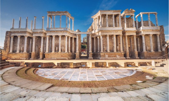 Vistas frontal del teatro romano de Mérida en la que se ve la fachada principal en todo su esplendor y el escenario. De fondo está el cielo azul con ráfagas de nubes que acarician el teatro. 