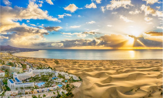 Vista panorámica del la playa de Maspalomas tomada durante el amanecer. En la imagen se pueden ver las edificaciones construidas junto a la inmensa playa de Maspalomas. Las gigantes dunas de arena blanca llegan hasta la orilla del mar totalmente en calma con diferentes tonos de azul sobre el que se reflejan los rayos del sol al amanecer. 