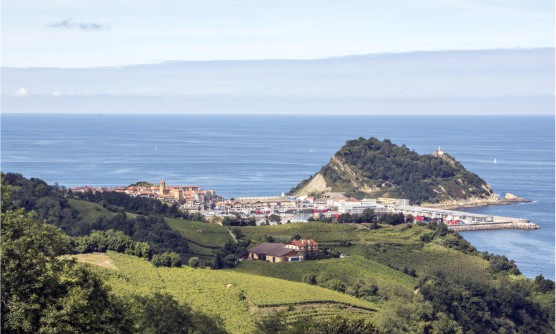 Vista panorámica de la ciudad de Zarautz tomada desde lo alto de una colina, con vistas al mar, la ciudad  y el mismo faro que también se encuentra sobre otra pequeña península. Las colinas están forradas de una verde pradera que lleva hasta la ciudad y hasta la orilla del mar.  