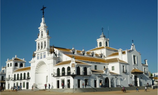 Imagen del Santuario de la Virgen del Rocío. Un edificio totalmente blanco situado en un cambio sin asfaltar. En el frontal del santuario se encuentra el campanario con tres campanas y un gran portón con el techo en forma de concha marina. El santuario está formado por diferentes dependencias a diferentes alturas, tejados de teja marrón, puertas de arco de medio punto y algunas ventanas del último piso circulares. Se ve grupos de gente charlando cerca de las escaleras que llevan al santuario y un jinete con su caballo.   