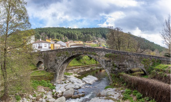 Puente de Aquelcabos, puente de piedra romano y medieval de Arenas de San Pedro. El Puente cruza el río Arenal con un caudal tranquilo, en el que únicamente varias rocas bajo el puente intentan cortar su paso. Al fondo se ven algunas viviendas con fachadas de color blanco, amarillo y salmón claro y tras ella una gran arboleda que cubre una colina. 