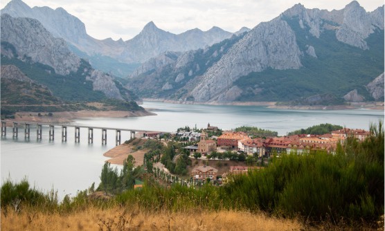 Vista panorámica del embalse de Riaño en la que se puede ver el pequeño pueblo a orillas del embalse y un gran puente que lo cruza. En el fondo de la imagen podemos ver las colosales montañas en un día nublado. 