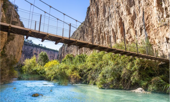 Puente colgante y peatonal de Chulilla sobre el río Turia. Las paredes de los acantilados que bordean el río son prácticamente verticales y rocosas, la vegetación se encuentra a la orilla de río caudaloso que se muestra en diferentes tonalidades de azul. 