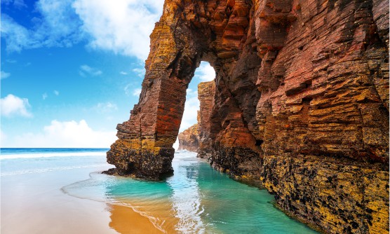 Hermosa imagen de la playa de las Catedrales en Ribadeo, provincia de Lugo. En la imagen se ve como la marea comienza a cubrir la arena de la playa. El mar tiene diferentes tonos de azul (azul marino al fondo y azul turquesa sobre la misma playa) en la que gigantes rocas en forma de arco se alzan como enormes monumentos naturales. Al fondo se ve como las olas golpean con fuerza la roca dejando un rastro de espuma blanca. 