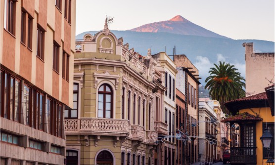 Imagen tomada desde La Orotava con el Teide al fondo con los últimos rayos de sol durante el anochecer bañando la cumbre. En la ciudad se ve un edificio señorial con balcones totalmente ornamentados. Frente a este otro edificio con un invernadero tallado en madera oscura. Al final de la calle hay dos grandes palmeras. 