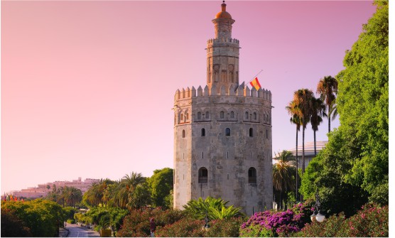 Imagen de la robusta Torre del Oro durante el atardecer. En primer plano se puede ver la Torre como protagonista rodeada de verdes arbustos, palmeras y flores en tonos fucsia. El cielo sin ninguna nube tiene tonos rojizos. 