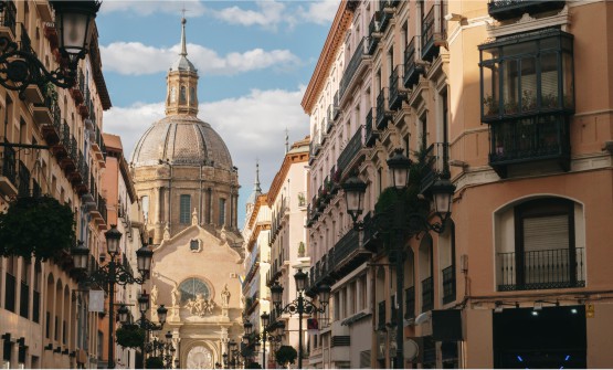 Calle peatonal de Alfonso I en la ciudad de Zaragoza, con edificios señoriales mayoritariamente con balcones de hierro forjado y fachadas de colores suaves. Al fondo se ve la magnífica cúpula de la basílica del Pilar. 