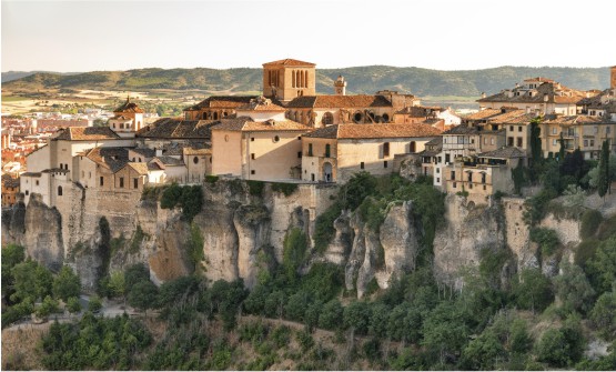Imagen tomada desde lo alto en la que se contemplan las casas colgantes de la ciudad de Cuenca situada sobre la cornisa rocosa. En el fondo de la imagen vemos una robusta torre con 4 ventanales verticales en cada lateral y al fondo una torre más estrecha con un reloj. 