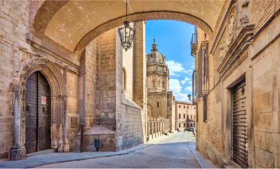 Calle del centro histórico de la ciudad de Toledo en la que se ve parte la cúpula de una iglesia. En primer plano vemos como las fachadas de ambos laterales de la calle están unidas por un pasadizo donde cuelga una lámpara de hierro forjado y cristal. 