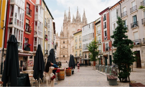 Calle peatonal de la ciudad de Burgos con algunas personas paseando. Se ven algunas terrazas de cafeterías con mesas y sillas, dos barriles que se usan a modo de mesa, diferentes contenedores para el reciclaje y 2 bancos de madera sin respaldo. Tiene edificios señoriales de colores intensos como el granate y amarillo, muchos de ellos con invernaderos o balcones. Al fondo de la imagen se ve parte de la catedral de Burgos y sobre todo sus altas agujas que parece que están rozando las nubes. 