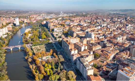 Vista aérea de la ciudad de Valladolid en la que se ve el río Pisuerga y del puente Juan de Austria. Una extensa zona verde separa el río de la ciudad y junto a esta una amplia avenida, de la que parte la zona residencial con grandes pisos, casas, plazas, iglesias, etc. Al fondo se puede ver la zona industrial y unas pequeñas colinas. 