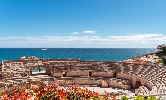 Vista panorámica del anfiteatro romano junto al mar Mediterráneo en Tarragona. En primer plano se ven las hojas verdes y rojizas de algunas plantas y detrás de ellas una vista parcial del Teatro. Al fondo de la imagen vemos el mar azul y con reflejos brillantes por los rayos del sol, con un pequeño oleaje y 4 grandes embarcaciones. 