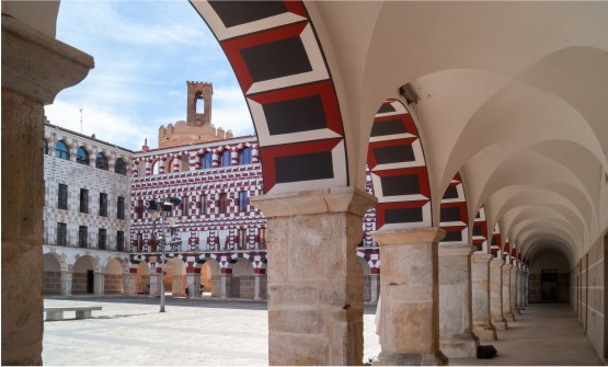 Imagen de la plaza alta de la ciudad de Badajoz, con la torre de los Espantaperros al fondo. En la imagen vemos una de las fachadas, así como bajo los arcos de la zona porticada, con grecas pintadas en rojo, negro y blanco.   