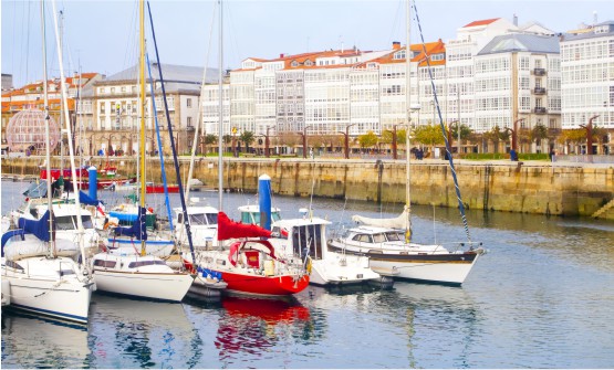 Imagen del puerto deportivo de la ciudad de A Coruña en la que se ven en primer plano varias embarcaciones de recreo amarradas en un mar de tonos plateados. Al fondo de la imagen, vemos un largo paseo con edificios con fachadas de vidrieras blancas. 