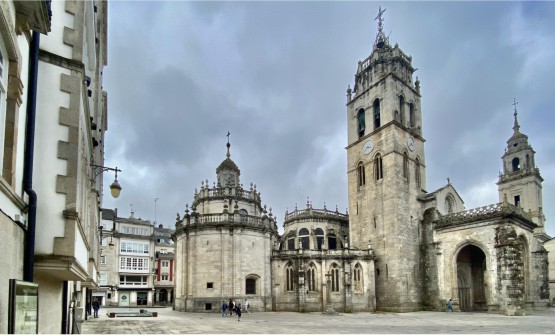 Imagen de la Catedral de Lugo del siglo XII Patrimonio de la Humanidad de la UNESCO tomada desde la plaza de Santa María en un día gris y totalmente cubierto por grandes nubes. De la catedral destaca en primer plano una de sus grandes torres con un reloj, así como sus naves laterales. 