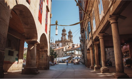 Vista de una calle peatonal en el casco antiguo de Pontevedra, con soportales a ambos lados de la calle y vistas hacia la iglesia de la Peregrina. En el fondo de la imagen también se ve un restaurante con un toldo blanco y personas sentadas en la terraza y paseando.  